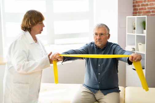 Ein älterer Mann sitzt auf einer Liege in einer Praxis, er macht eine Übung mit einem gelben Thera-Band. Eine ältere Frau im weißen Kittel, die wie eine Physiotherapeutin aussieht, spricht mit ihm. 