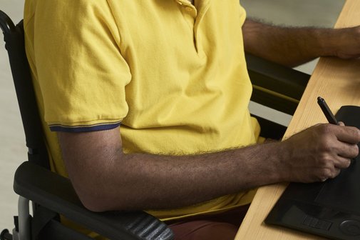 Ein Mann im Rollstuhl sitzt an einem Schreibtisch, zeichnet etwas auf einem Grafik-Tablet. 
